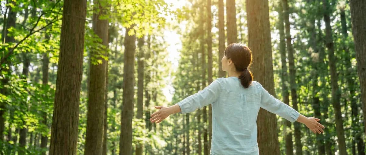 A woman standing in a sunlit forest with her arms outstretched